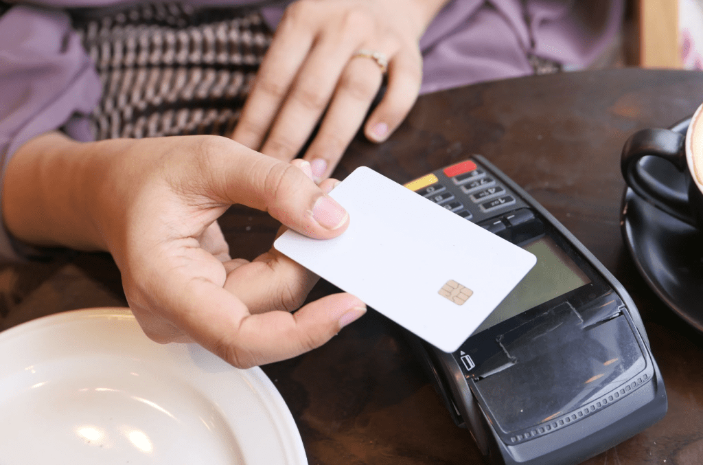 A person's hand taps a payment card to a payment terminal on a table for contactless payment.