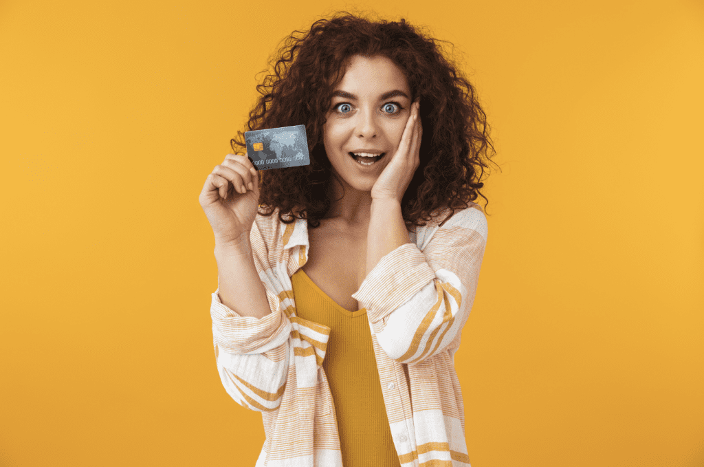 A woman with dark, curly hair stands in front of a yellow background and is holding up a credit card in one hand and holding the other hand to her face with an expression of pleasant surprise.