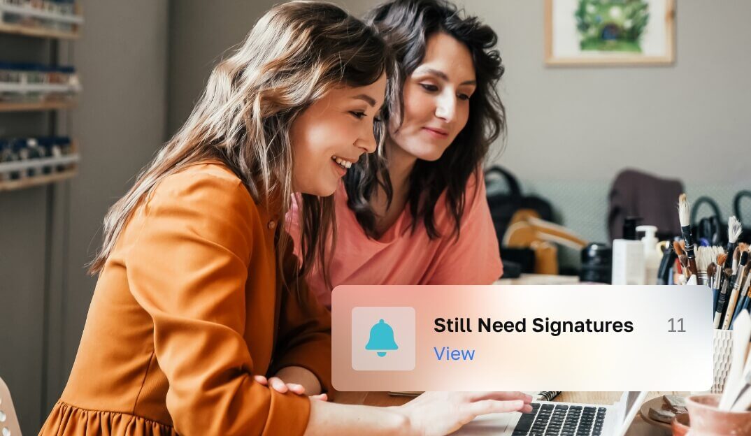 Two women dressed in orange and pink blouses, sitting on a laptop browsing an Internet site.